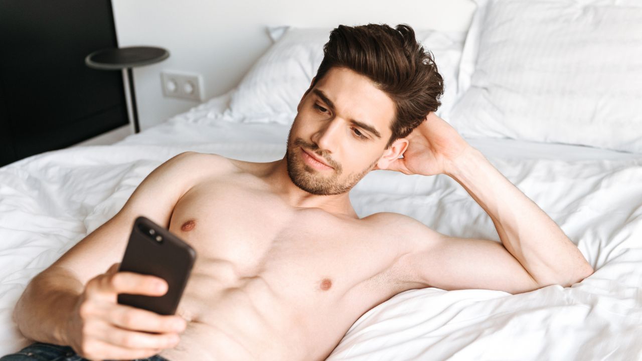 A shirtless man lying on a bed, holding and looking at a smartphone.